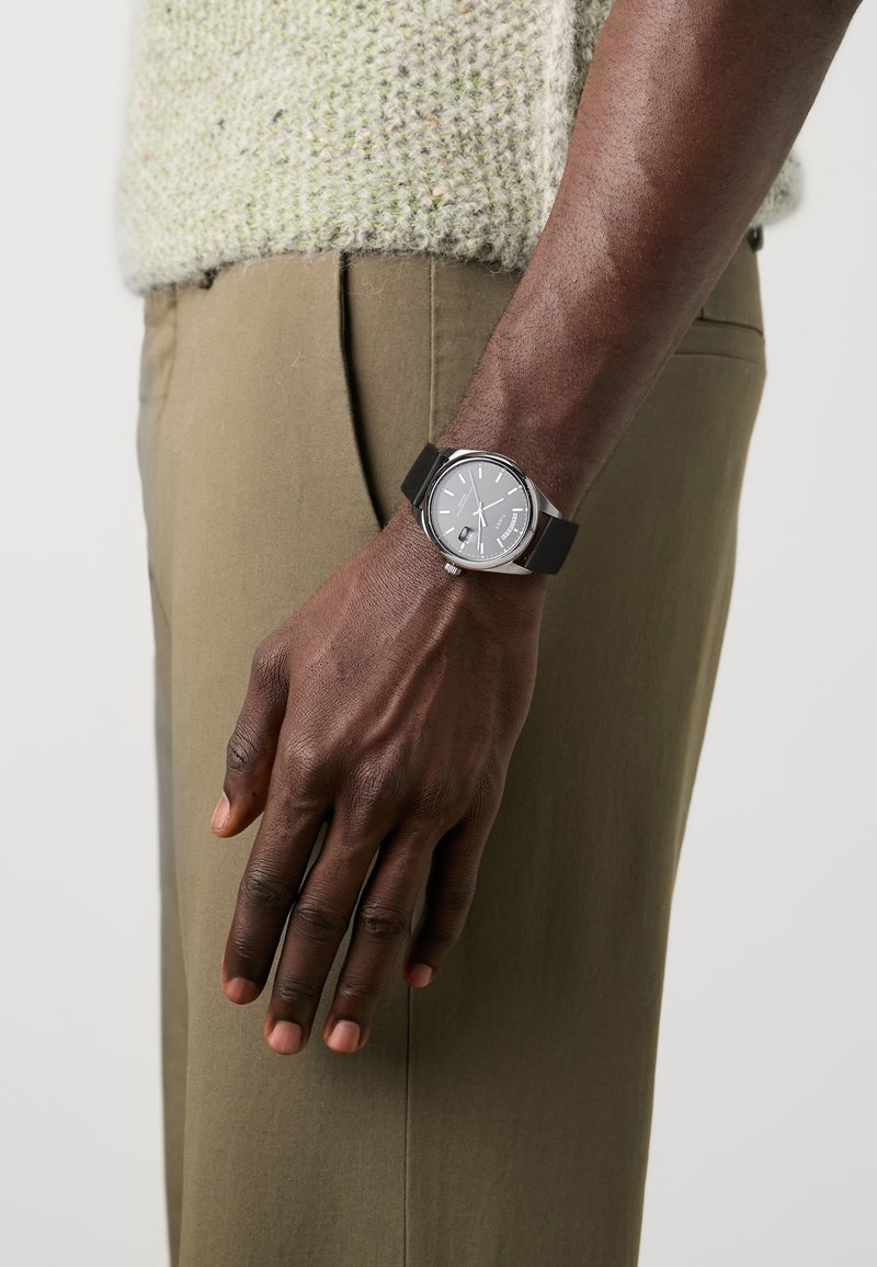 Montre en argent avec un cadran gris, un bracelet en caoutchouc noir et des accents argentés. Portée sur une main sur un tissu de pantalon vert olive.