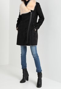 Cappotto di suede nero con un ampio colletto in pelliccia sintetica chiara, zip frontale asimmetrica, maniche lunghe e tasche laterali, abbinato a jeans blu.
