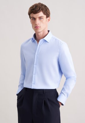 Młody mężczyzna w jasnoniebieskiej, długorękawowej koszuli zapinanej na guziki, włożonej do czarnych plisowanych spodni, stojący z rękami w kieszeniach.