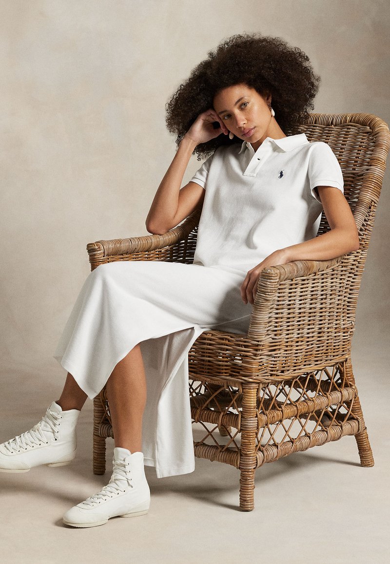 Valkoinen poolopaita, jossa pieni logo, yhdistettynä pitkään valkoiseen hameeseen. Malli istuu punotussa rottinkituolissa ja käyttää valkoisia korkeavartisia lenkkareita.