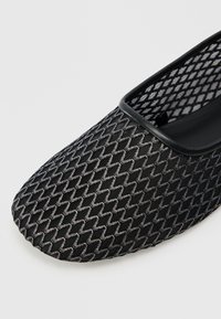 Scarpa nera slip-on con tomaia in mesh testurizzata caratterizzata da un motivo a zigzag e punta arrotondata e liscia. Un bordo nero delimita l'apertura.