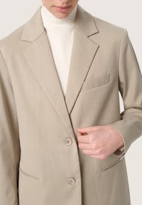 Beige blézer textúrált felülettel, két gombbal és zsebbel. Fehér bordázott garbó fölött viselve, amely a szabott dizájnját hangsúlyozza.