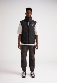 Svart vadderad väst med hög krage, frontdragkedja och logopatch. Buren över en vit skjorta, tillsammans med mörka cargobyxor och sneakers.