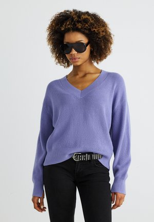 Femme aux cheveux bouclés portant de grandes lunettes de soleil noires, un pull col en V lavande, un pantalon noir et une ceinture cloutée.