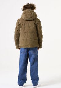 Olivgrüner Cord-Puffermantel mit Kapuze, Rückansicht, mit gesteppten Abschnitten. Getragen mit blauen weitgeschnittenen Jeans und weißen Turnschuhen.