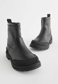 Bottines noires en cuir synthétique lisse avec une semelle en caoutchouc texturé, dotées d'une fermeture éclair latérale et d'une languette pour un enfilage facile.