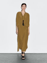 Robe vert olive avec un décolleté en V profond, des manches bouffantes et une taille cintrée. Associée à des sandales noires à lanières et un long collier à pendentif.