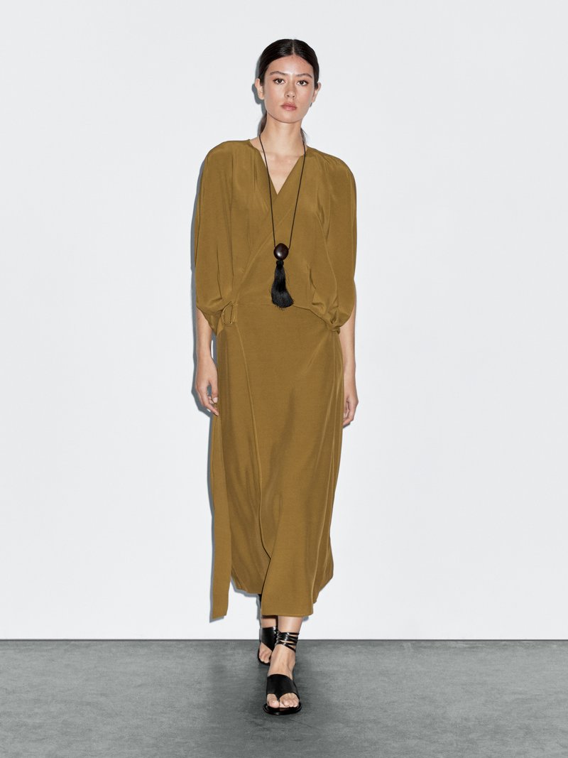 Robe vert olive avec un décolleté en V profond, des manches bouffantes et une taille cintrée. Associée à des sandales noires à lanières et un long collier à pendentif.