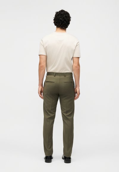 Άνδρας με σγουρά μαλλιά στέκεται με την πλάτη, φοράει μπεζ κοντομάνικο πουκάμισο, ελαιοπράσινο παντελόνι και μαύρα παπούτσια με απλό φόντο.