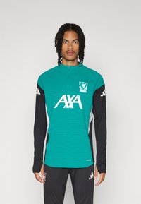 Žalias zip-up treniruočių viršus su juodomis ilgomis rankovėmis ir baltais akcentais. Priekyje yra Liverpulio FC logotipas ir „AXA“ ženklinimas.