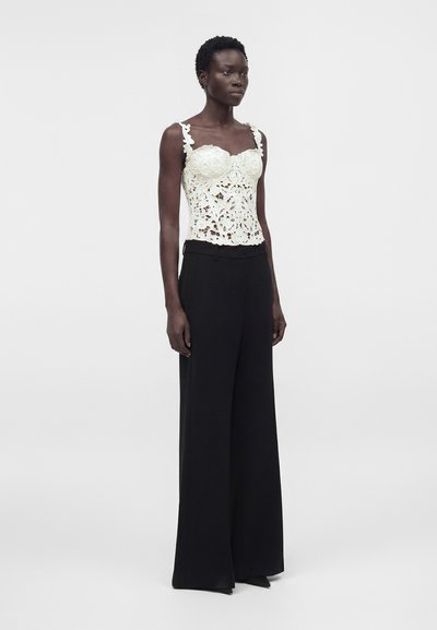 Λεπτή γυναίκα που φοράει λευκό λουλουδάτο δαντελένιο αμάνικο τοπ και ψηλόμεσα μαύρα παντελόνια με φαρδιά πόδια, στέκεται μπροστά σε απλό ανοιχτό φόντο.