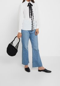 Blusa bianca con bordo in pizzo nero e fiocco al colletto, abbinata a jeans blu a zampa e mocassini neri con texture. Borsa nera intrecciata.