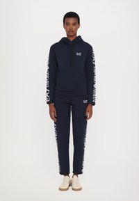 Sudadera con capucha navy y joggers a juego hechos de material suave. Diseño con capucha y bolsillo delantero, y acentos de logo blanco en las mangas y las piernas.