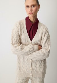 Creme kabelgebreide cardigan met een V-hals, grote knopen en geribde manchetten, gecombineerd met een burgundy overhemd met kraag daaronder.