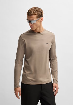 Beige Langarmshirt mit Rundhalsausschnitt, subtilen Logo auf der Brust und glatter Stoffoberfläche. Getragen mit grauen Sonnenbrillen.
