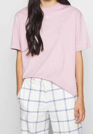 Persoon die een lichtroze Nike t-shirt en witte broek met blauw geruit motief draagt, staand met één hand in de zak tegen een effen achtergrond.