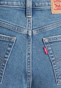 Jeans en denim bleu clair avec un design de couture orange, dotés de deux poches arrière et d'une étiquette rouge sur la poche droite.