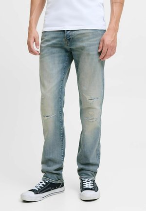 Man wearing faded blue ripped jeans, white t-shirt, and black sneakers with white laces standing on a white background.