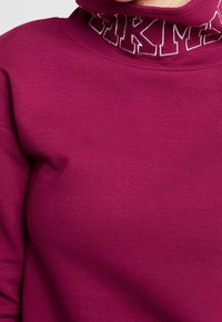 Vínová mikina s vysokým límcem a stříbrným vyšívaným nápisem v kontrastní barvě. Jemná, hladká textura bez vzorů.