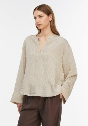 Kobieta ubrana w beżową, prążkowaną bluzkę z długim rękawem, dekoltem w serek i szerokimi mankietami, w połączeniu z luźnymi, ciemnobrązowymi spodniami, stojąca na białym tle.