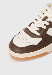 Baskets marron et beige avec une tige en cuir lisse, boîte de talon perforée et lacets blancs. La semelle est de couleur gomme et légèrement texturée.