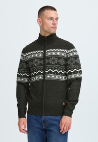 Mørkegrønn strikket genser med høy krage som har fair isle-mønster i hvitt og grått over brystet. Med glidelås foran og ribbestrikkede mansjetter.