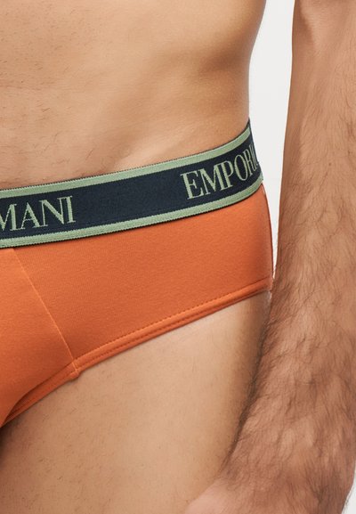 Torso et bras masculins portant un slip orange avec une ceinture noire et verte affichant le logo Emporio Armani.