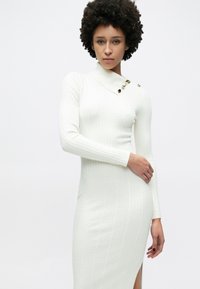 Μακρύ λευκό πλεκτό φόρεμα με ανάγλυφη υφή, που διαθέτει ψηλή λαιμόκοψη και χρυσά κουμπιά σε έναν ώμο. Διαθέτει πλαϊνή σχισμή.