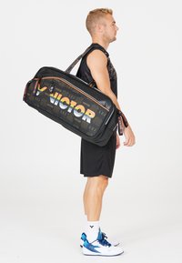 Sort sportstaske i duffel stil med orange og hvit "VICTOR"-logo, geometriske mønstre, justerbar strop og slidstærkt materiale.