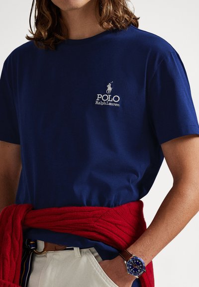 Μπλε βαμβακερό μπλουζάκι με κοντά μανίκια, διακοσμημένο με κεντητό λευκό λογότυπο και κείμενο. Κόκκινο πλεκτό πουλόβερ δεμένο στη μέση.