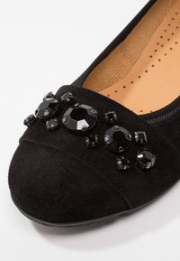 Chaussure plate en suede noir avec des ornements en gemmes facettées noires sur le bout et une semelle intérieure perforée beige.