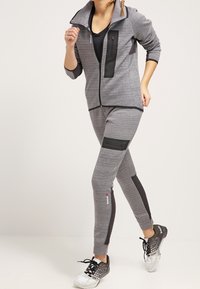 Giacca sportiva grigia con accenti neri e tasca con zip, abbinata a pantaloni grigi con pannello nero, indossata con scarpe sportive.
