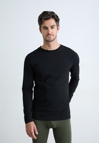 Svart langermet t-skjorte med rund hals, glatt tekstur, tettsittende design, brukt sammen med olivengrønne leggings mot en ensfarget bakgrunn.