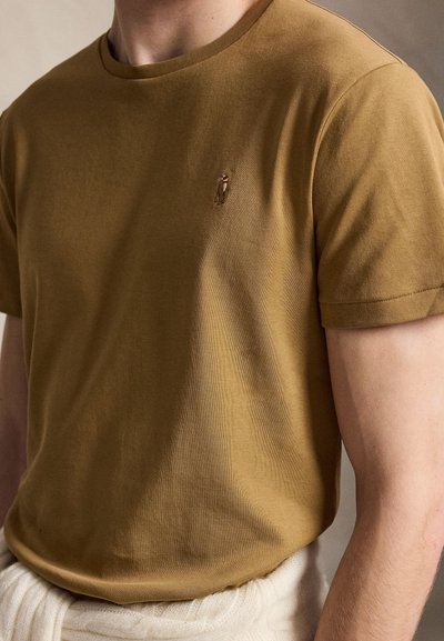 Άνδρας που φοράει κοντομάνικο t-shirt σε μουσταρδί καφέ χρώμα με μικρό κεντημένο λογότυπο στο στήθος, μπεζ πλεκτό δεμένο γύρω από τη μέση.