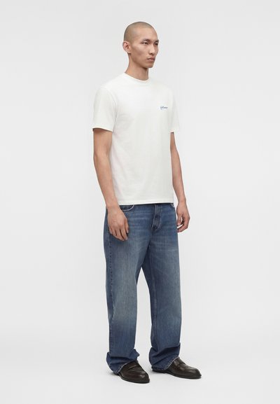 Άνδρας μοντέλο στέκεται μπροστά φορώντας λευκό κοντομάνικο μπλουζάκι με μικρό μπλε λογότυπο και χαλαρά μπλε τζιν με μαύρα παπούτσια.