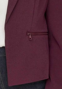 Blazer bordeaux avec une texture lisse, doté d'une poche zippée sur le côté et d'une coupe structurée, associé à un top blanc et un denim foncé.