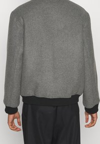 Person som bär en grå ulljacka med svarta manschetter och midjeresår, visad bakifrån, tillsammans med löst sittande svarta byxor.