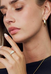 Guldörhänge, glittrande guldring med en päronformad design, mjuk hudton och neutral nagellack mot en mörk bakgrund.