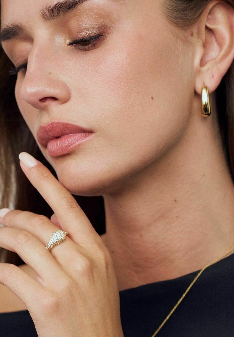 Guldörhänge, glittrande guldring med en päronformad design, mjuk hudton och neutral nagellack mot en mörk bakgrund.