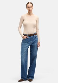 Beżowa bluzka z długim rękawem i dekoltem w serek, zestawiona z luźnymi niebieskimi jeansami i brązowym paskiem. Całość uzupełniają ciemnobrązowe buty.