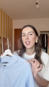 Hosszú sötét hajú nő arany fülbevalókkal, aki egy világoskék inget tart egy fehér akasztón egy hálószobai szekrényben.