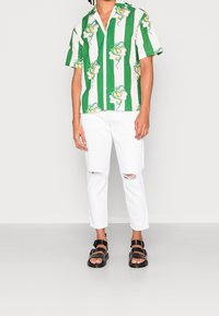 Zeleno-bílé pruhované tričko s krátkým rukávem a květinovým vzorem, v kombinaci s bílými potrhanými džínami a černými sandály na pásky.