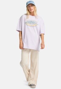 Camiseta oversized de color púrpura claro con un estampado gráfico, combinada con pantalones anchos de canalé beige y una gorra azul clara. Zapatillas grises visibles.