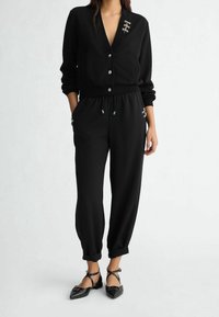 Kobieta ubrana w czarny kardigan zapinany na guziki ze srebrnymi zapięciami, czarne spodnie ze ściągaczem oraz czarne spiczaste baleriny z paskiem wokół kostki.