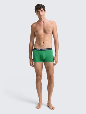Mužský model stojící bosý, oblečený v zelených boxerkách s černým páskem s nápisem "Tom Tailor" na pozadí jednoduchého světlého pozadí.