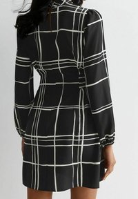 Abito nero con motivo a griglia bianca, maniche lunghe, vita aderente e spalle a sbuffo, con apertura sul retro e tessuto dalla texture liscia.