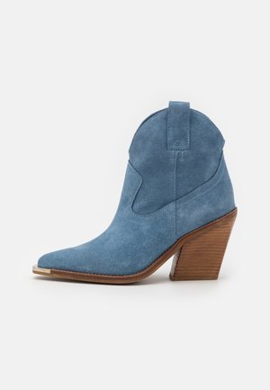 Bottine en daim bleu avec bout pointu, talon bloc en bois empilé et languette à l'arrière, sur fond blanc.