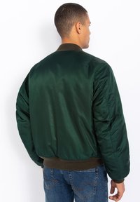 Giacca bomber verde in satin con colletto e orlo a costine, caratterizzata da spalle imbottite e una texture liscia. La vista posteriore mette in mostra le linee di design.