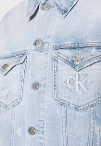 Giacca di jeans blu chiaro con colletto classico, due tasche sul petto, bottoni argento e logo "CK" ricamato sulla tasca sinistra.
