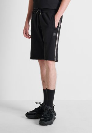 Man die zwarte sportieve shorts met witte zijstrepen draagt, zwarte sokken en zwarte sneakers, staat met één hand in de zak.